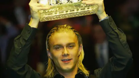 Getty Images The snooker player Paul Hunter, lifting a trophy aloft out of shot. His blond hair is braided and he is wearing a black shirt.