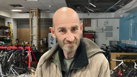 Grace Wood/BBC A man wearing a puffer cord jacket stands in a bike warehouse looking down the camera.