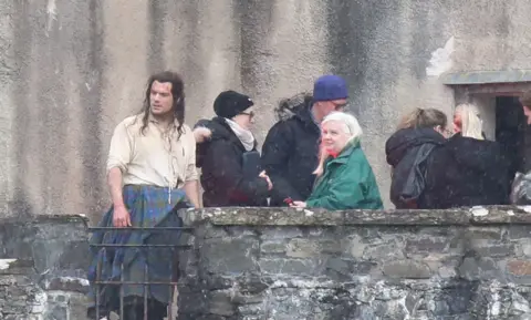 Peter Jolly Henry Cavill with long hair and wearing a kilt and an off-shirt. He is next to castle and standing next to a group of people wearing warm coats and hats. 