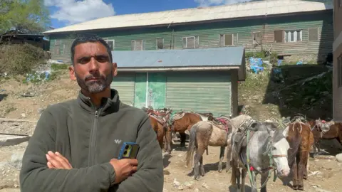 A man in a green fleece jacket stands in the foreground with his arms crossed. To the right, are several horses and behind him are two green sheds. 