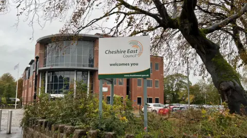 BBC A view of Cheshire East Council's former headquarters at Westfields in Sandbach. There is a sign in some bushes saying "Cheshire East Council welcomes you". Behind it is a red brick building with a curved glass frontage in front of a car park.