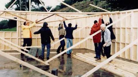 Kendal Snowsports Club A group of people work together to hold timber up on a site which is half-built. They are wearing dated clothes from the 1980s and overalls because it looks like it is raining with wet conditions. There are planks of timber holding the facility up. 