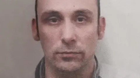 Daniel Washbourne is looking at the camera with a stubbly moustache. He has short brown hair. The image is low-quality. 