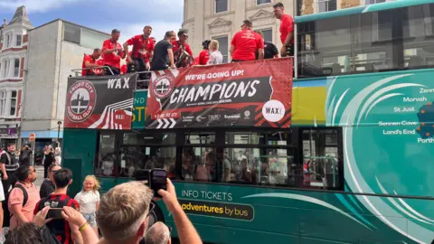 Players are on the top deck of the open bus and it is in the middle of the city. There are spectators looking up and taking photos. A banner on the bus reads: WE'RE GOING UP. CHAMPIONS
