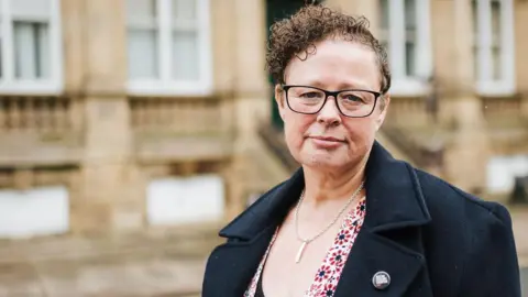 Local Democracy Reporting Service Sarah Courtney wears glasses a dark coat and a flowery top as she looks directly at the camera. In the background is a blurred building.