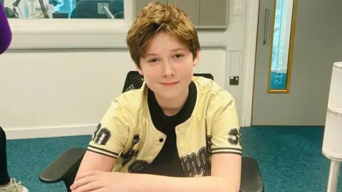 Albie Salter is sitting in an office chair in the Radio Berkshire Studio, he is smiling at the camera, wearing a black t-shirt and a yellow shirt over the top.