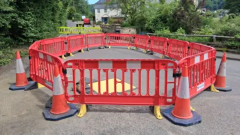 A large circular arrangement of cones and bollards blocks a quiet leafy road. 