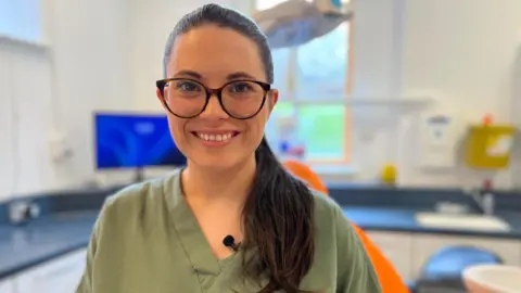 Katie Davis smiles at the camera. She has long, dark hair and is wearing dark-framed glasses. Behind her is a dentist's clinic space. It is a head-and-shoulders shot; she is in focus while the background is blurred.