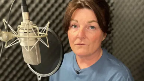Gayanne sits in front of a microphone in a grey soundproofed recording booth, she has brown hair and wears a blue T-shirt