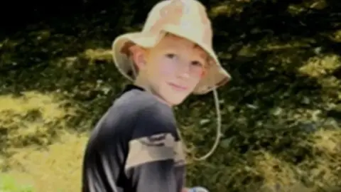 Zac Roe looking over his shoulder at the camera. He is wearing a dark top and a floppy summer hat. The background shows tree shadows on grass.