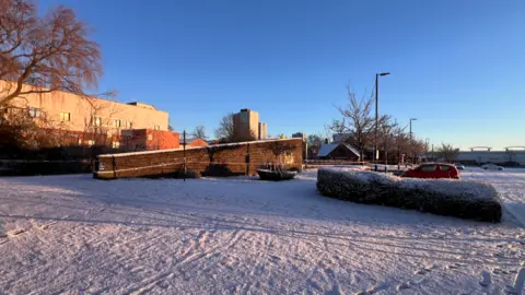 A snow covered area leading up to a bridge. There is red and white tape running from the bridge across towards a car park