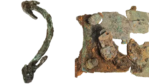 Anna Giecco The lockbox and its handle are laid out side by side. On the left, the curved piece of metal is discoloured green and has an arrow-head shaped ends. On the right, the severely-damaged bronze box is coloured grey and rusty red. It does not look like a box as it is photographed from above.