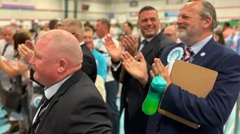 Martin Heath/BBC Three men in dark suits clap and look on. They are standing in a sports hall.