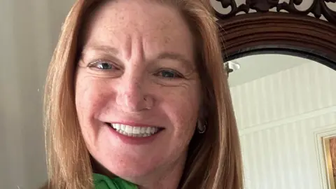 Wendi Merrit A woman with strawberry blonde hair smiles into the camera. She is standing in front of a mirror in a hotel room