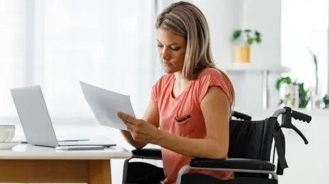 A woman sitting in a wheelchair in front of her laptop, looking at paperwork.