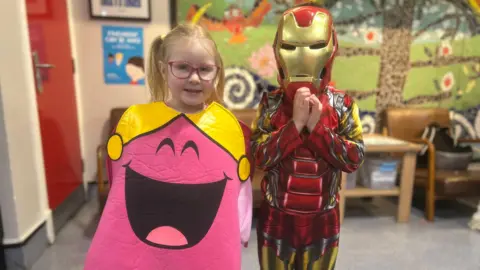 Isabel and Dylan. Isabel has blonde hair in pigtails, wearing pink glasses and a pink Miss Chatterbox costume. Dylan is dressed as Ironman.