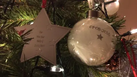 Ellie Goan A close up image of a beige gold star and a Christmas bauble on a Christmas tree. Lights and some other decorations can be seen on the tree. 'Our wee star Baby G is written 25/11/2015' is inscribed on the star.