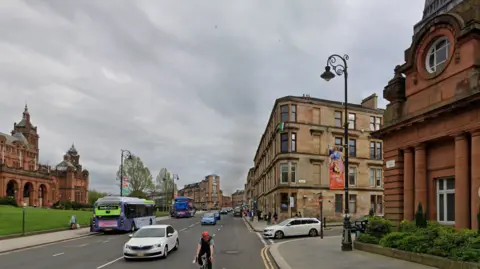 A blonde sandstone tenement building at the corner of Blantyre Street stands between the red sandstone Kelvingrove Museum and Galleries on the other side of the road, beyond a grassy bank and Kelvin Hall in the foreground.