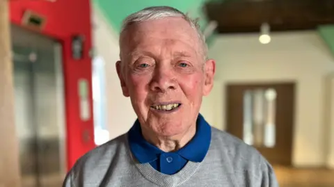 Close-up portrait of a 90-year-old Iain with short grey hair, wearing a grey sweater over a blue collared shirt, standing indoors with a blurred background featuring a red wall and doorway.