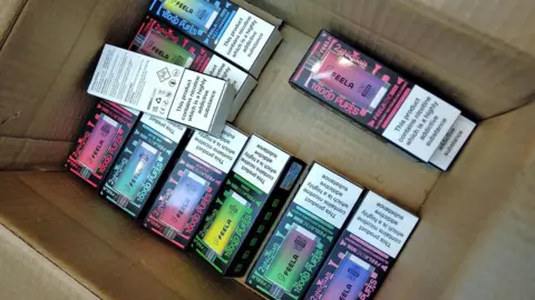 A cardboard box with 10 illegal vape boxes lying in the bottom. They are colourful designs with a picture of the rectangular vape on the front, along with a warning that the product is highly addictive.