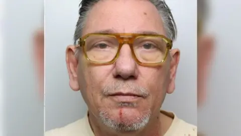 Thames Valley Police A police custody photo of a man with grey hair and grey stubble, wearing a pair of yellow glasses and a pale T-shirt. He's looking straight at the camera and has a plain white wall behind him.