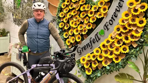 Stephen Aslin, wearing a white helmet and blue body-warmer, poses with his bike in front of a floral Grenfell memorial