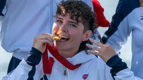 Lauren Rowles has brown hair and is wearing a white Paralympics jumper. She has a gold medal around her neck, which she is biting and smiling to the camera.