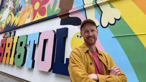 A photo of a man wearing a bright coloured yellow jacket and pink shirt, with his colourful artwork in the back