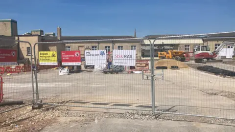 Chippenham Rail Station, with a building sire in front - large metal gates with piles of materials and machinery behind. Sunnny day.