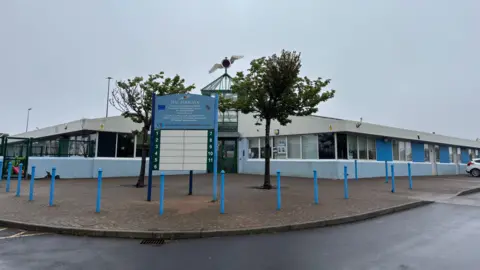 The phoenix community centre. It is a large, single storey building with a pitched roof, blue and white facade, and two trees planted out the front