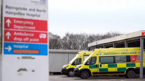 Two ambulances sit outside a hospital Accident and Emergency Department. An out of focus hospital sign can be seen in the foreground directing towards emergency departments.