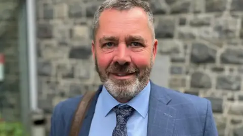 Man centre of image wearing a blue checkered blazer and a tie.