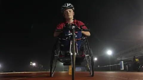 John Fairhall/BBC Morgan is pictured in his racing wheelchair on the athletics track. He is looking down at the camera with a determined expression on his face. He is wearing a silver helmet and a red t-shirt with racing gloves on his hand.