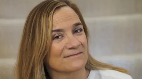 Tracy Chevalier has long blonde hair and is wearing a white shirt and smiling at the camera.