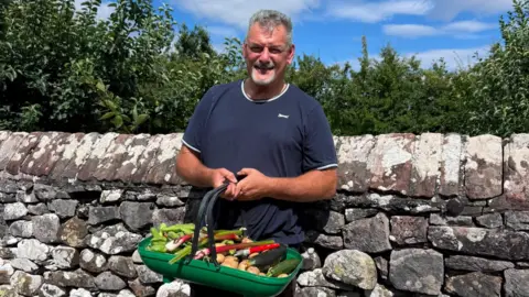 Adam Day Adam Day, a man with grey hair and a beard, standing against a dry stone wall holding a basket containing vegetables from his allotment in Greystoke. He is wearing a dark t-shirt and smiling at the camera. 