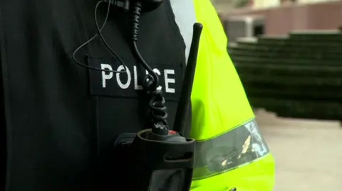 A closeup of a police officer's uniform. The hi-visibility sleeve can be seen, as can a radio in a pocket. Police is written just above the pocket.
