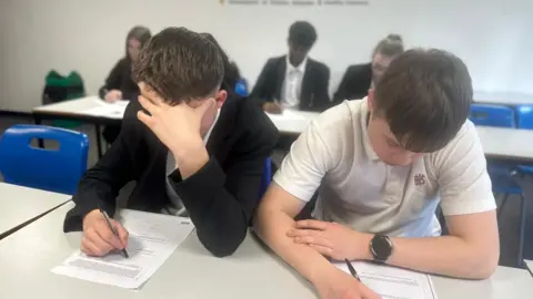 BBC Two boys, in black and white school uniform, sat at a white desk leaning over exam papers. Both their heads are down and faces not visible, and they both have brown hair. One has his head in his hands and they are both holding pens in their right hands. Behind them are more students, lined up sat a desk too.