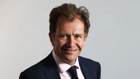 Thames Valley ICB Dr Nick Broughton smiles for a professional headshot photo. He is wearing a dark jacket and tie with a pink shirt, and has mid-length wavey dark hair