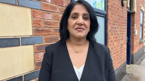 Woman with dark hair standing in front of a brick wall. She is wearing a white top and a black blazer and has a gold necklace on