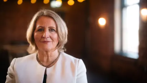 A portrait photo of a middle aged woman. She is smartly dressed in a white jacket. She has short, blonde, bobbed hair.