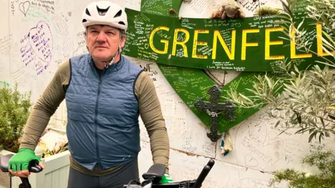 Stephen Aslin Stephen Aslin standing in front of the Grenfell Tower memorial. He is wearing a blue gilet over a brown top, green cycling gloves and a white cycling helmet. 