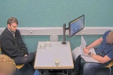 Essex Police CCTV still image of Brady sitting on one side of an interview desk. He is wearing a dark fleeced jacket, dark trousers and has his right leg crossed on his left knee. There are drinking cups on the table. On the other side is a police worker wearing a grey-blue polo shirt and a lanyard. His face, which is blurred, is to the side as he looks through a folder with plastic wallets of information. There is a computer screen bracketed to the table facing him. The walls behind the two men are a padded duck-egg blue