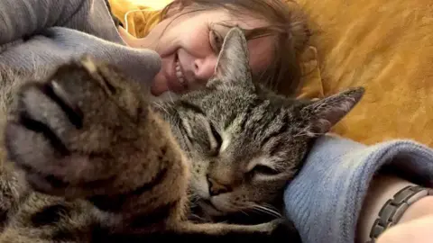 Lisa Burtenshaw Lisa Burtenshaw is pictured lying down with a tabby cat. The cat is resting its head on her arm and one of its paws is stretched out near the camera. Lisa is smiling and looking directly at the camera.