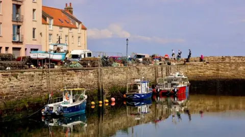 Graham Christie A row of brightly painted three fishing boats at a harbour. Several people can be seen walking around in the background of the harbour.