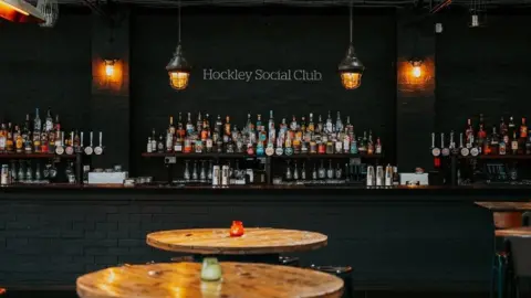 Hockley Social Club A bar with many different types of bottles in view with the sign Hockey Social Club above it. Several tables which each have a candle on can also be seen.