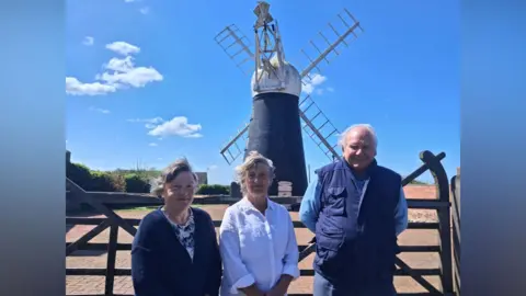 Two women and a man standing in front of a windmill. The windmill is black and has four main sails and a fantail to the rear.