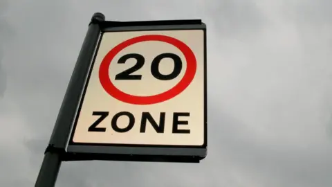 A 20mph zone sign on a pole with a grey sky as a back drop.