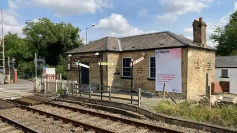 A small stone brick building sits behind railway lines on a sunny day. There's a railway crossing to the right with trees. 