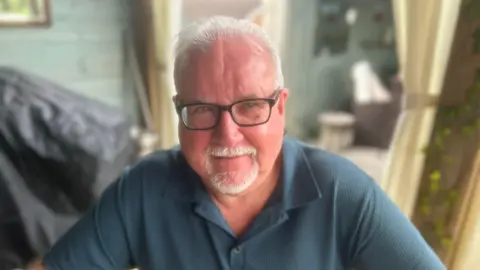 Philip Renshaw. He has white hair and goatee, wearing glasses and a blue polo top.