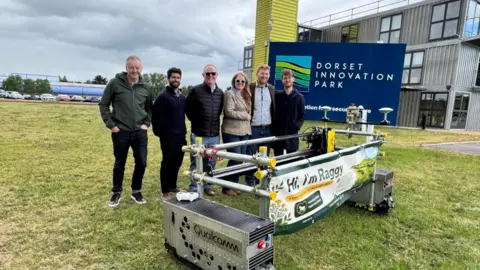 Six people stood behind a robot called Raggy on a field in front of a sign saying Dorset Innovation Park